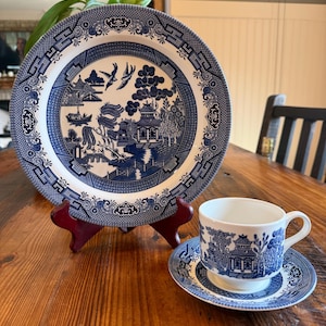 May include: A blue and white Willow pattern plate, teacup, and saucer set. The plate is displayed on a red stand. The teacup and saucer are on a wooden table. The design features a scenic landscape with pagodas and birds.