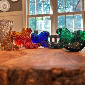 May include: A collection of decorative glass cowboy boot figurines in various colors, including amber, red, blue, and green. The boots are arranged on a wooden surface, showcasing their intricate designs and textures.