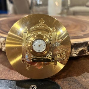 May include: A gold-toned compact with an embedded watch. The watch face is white with black numerals and hands. The compact features an ornate design with pillars and a crown. The compact is round and appears to be made of metal.