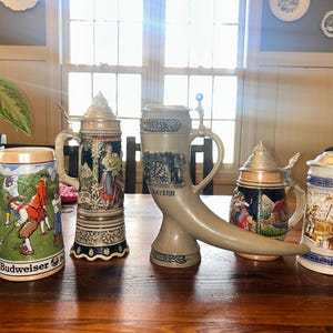 May include: Collection of decorative beer steins with lids, featuring various designs and scenes. The steins are made of ceramic and glass, with handles and metal lids. One stein is shaped like a horn.