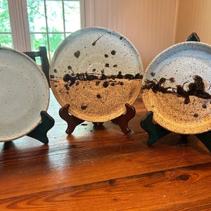 May include: Three ceramic plates with unique designs. The plates are displayed on wooden stands. The plates have a speckled, light blue and brown color scheme with dark brown paint splatters. The plates are on a wooden table.