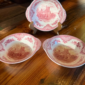 May include: Three white ceramic bowls with pink transferware patterns featuring a castle landscape in the center and floral motifs along the scalloped rims. Each bowl has two small side handles and a glossy finish, displaying a consistent design.