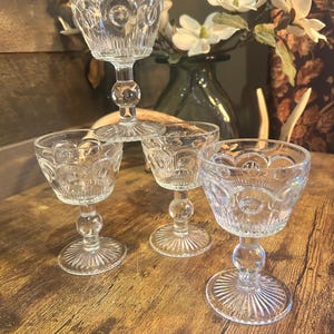 May include: Four clear, vintage-style stemmed glasses with detailed patterns. The glasses have a round bowl, a decorative stem, and a circular base. They are arranged on a wooden surface, with a vase of flowers in the background.