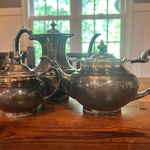 May include: Antique silver tea set with a teapot, creamer, and sugar bowl. The teapot has a curved spout and ornate handle. The creamer and sugar bowl have matching designs. The set is displayed on a wooden table.