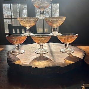 May include: A pyramid of six amber-colored, ribbed glass dessert or champagne glasses. The glasses are arranged on a wooden slab, with sunlight streaming through a window in the background. The glasses have a classic, vintage design.