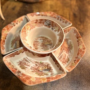 May include: A vintage ceramic serving dish set with a central bowl and four surrounding sections. The dishes feature a detailed landscape scene in shades of brown, orange, and cream, with floral accents around the rims. The set is designed for serving snacks or appetizers.