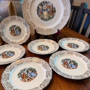 May include: A collection of vintage white porcelain plates with gold filigree rims. Each plate features a central image of a couple in period clothing. The set includes various sizes, from large serving plates to smaller dessert plates.