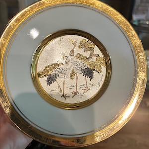 May include: A decorative plate with a gold rim and a light blue border. The center features a detailed illustration of two cranes and a tree in gold, black, and white. The plate has a gold rim and a light blue border.