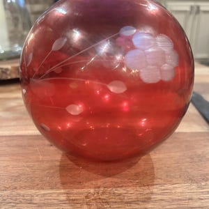 May include: A round, red glass vase with etched floral and leaf designs. The vase has a spherical shape and sits on a wooden surface. The etched designs are white and create a contrast against the red glass.