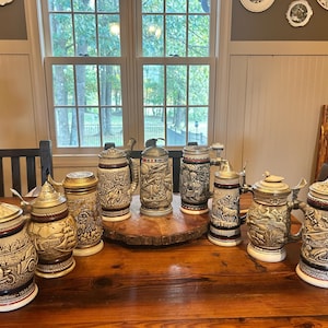 May include: A collection of vintage beer steins with detailed relief designs, including scenes of people, animals, and historical events. The steins have metal lids and handles, and are displayed on a wooden table.