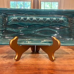 May include: A rectangular teal ceramic serving dish with a textured wave design. The dish is supported by a gold-colored stand. The dish has a central recessed area with a series of small, raised circles.