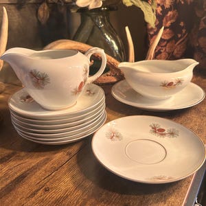 May include: A vintage white porcelain gravy boat, creamer, and saucers set. The set features a gold rim and a repeating pattern of red and green pine cones. The creamer and gravy boat are on top of a stack of saucers.