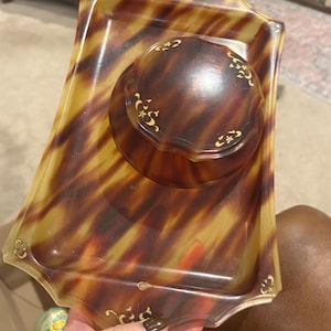May include: A vintage vanity set featuring a rectangular tray and a round lidded container. The set has a tortoiseshell pattern in shades of brown and yellow, with gold-colored decorative accents. The tray measures approximately 8 inches by 6 inches.