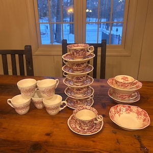 May include: A collection of vintage red and white teacups, saucers, and a serving bowl are arranged on a wooden table. The teacups and saucers are stacked in a tower, showcasing a detailed pattern. The bowl is oval-shaped with a matching design.