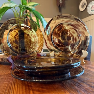 May include: A set of amber-colored glass plates with a spotted pattern. The plates are stacked on a wooden table, with two larger plates displayed on stands in the background. The plates have a warm, inviting aesthetic.