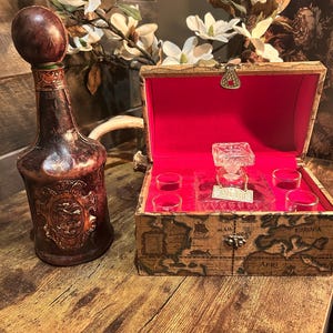 May include: A vintage liquor decanter with a dark brown leather-like exterior and a large, round stopper. Beside it is a wooden box lined with red velvet, containing a glass decanter and four shot glasses. The box is decorated with a map design.