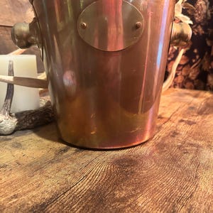 May include: A copper-colored ice bucket with brass handles and an oval brass plate. The bucket has a cylindrical shape and sits on a wooden surface. The handles are rounded and attached to the sides of the bucket.