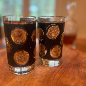 May include: Two clear highball glasses with a black band adorned with gold-colored coin-like embellishments. The coins have text and are set against a textured background. The glasses sit on a wooden surface.
