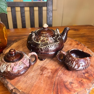 May include: A brown ceramic tea set with a teapot, sugar bowl, and creamer. The teapot has a lid and spout, and the sugar bowl has a lid and handles. The creamer has a handle and a small spout. The set is on a wooden surface.