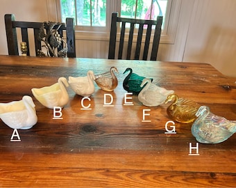 May include: A collection of decorative swan figurines in various colors, including white, yellow, clear, amber, green, and iridescent blue. The swans are arranged on a wooden table, showcasing their different designs and textures.
