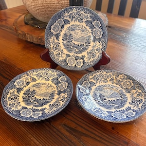 May include: Three blue and white decorative plates with a floral and landscape design. The plates are displayed on a wooden table. The plates have a detailed border and a central image of a landscape with trees and mountains.