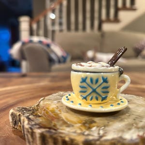 May include: A miniature yellow ceramic teacup and saucer set with blue snowflake designs. The cup is filled with a white substance and topped with a small brown stick. The set is placed on a wooden surface.