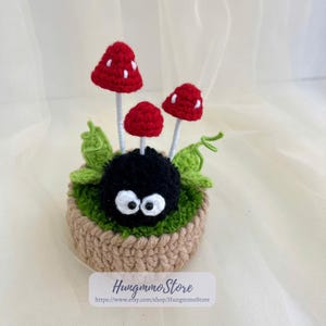 May include: A crocheted decorative piece featuring a black creature with white eyes, surrounded by green leaves and red mushrooms with white spots. The piece is set in a brown crocheted pot. The text "Hungmmo Store" and a website address are visible.
