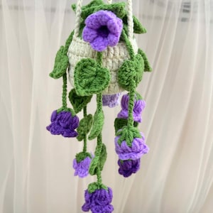 May include: A handmade crochet hanging planter featuring a cream-colored pot with green leaves and purple flowers. The planter is suspended by a cream-colored rope. The flowers are a vibrant purple, and the leaves are a deep green.