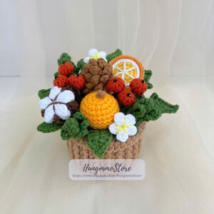 May include: A crocheted arrangement of various fruits and flowers in a woven basket. The arrangement includes an orange slice, a pumpkin, red berries, white flowers, and green leaves. The basket is tan, and the overall design is handmade.