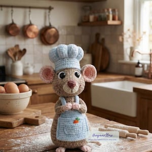 May include: A crocheted mouse figurine wearing a chef's hat and apron, standing on a wooden surface. The mouse has large ears, a pink nose, and is holding its hands together. The apron has a small carrot applique.