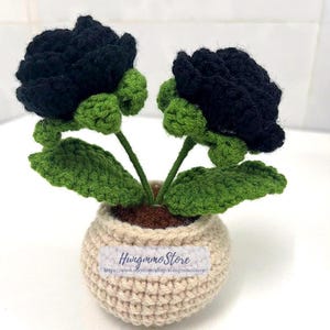 May include: A crocheted flower arrangement featuring two black roses with green leaves and stems, set in a beige pot. The pot has a label with the text "Hungmmo Store" and a website address.