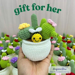 May include: A crocheted white pot with a smiling yellow bee and green cactus plants with yellow and pink flowers. The text "gift for her" is in green. The background features more crocheted cactus plants.