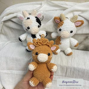 May include: Three crocheted cow keychains in white, brown, and black. The keychains have small black eyes, pink ears, and white hooves. The keychains are handmade and have a loop for attaching to keys or bags. The keychains are soft and plush.