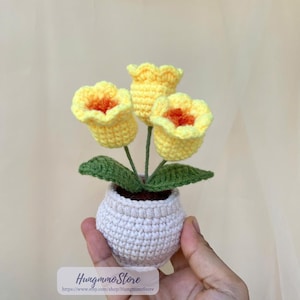 May include: A crocheted flower arrangement featuring three yellow tulips with orange centers, green leaves, and stems, presented in a small, off-white crocheted pot. The arrangement is handmade and decorative.