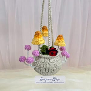 May include: A handmade crochet hanging planter featuring a gray pot with a mushroom and ladybug design. The mushrooms have yellow and orange caps. The planter is suspended by a braided cord. The text "HungmmoStore" is visible at the bottom.
