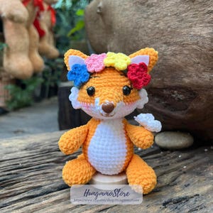 May include: A crocheted fox doll with orange fur, white belly, and a colorful floral crown. The fox has black eyes and a brown nose. The doll is sitting on a wooden surface. The text "Hungmmo Store" and a website address are visible.