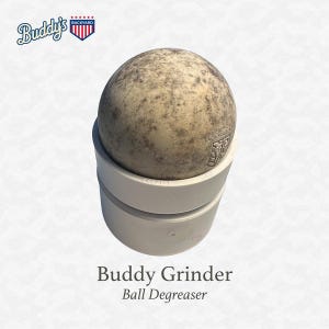 Puede incluir: Se muestra un desengrasante de bolas de lacrosse, el "Buddy Grinder". La bola es de color marr&oacute;n moteado y se asienta sobre una base de pl&aacute;stico blanco. El texto "Buddy Grinder" y "Ball Degreaser" est&aacute;n debajo de la base. El logotipo de "Buddy's Backyard" est&aacute; arriba a la izquierda.