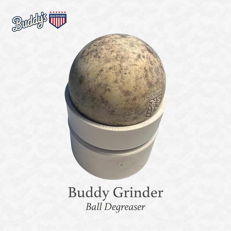 Puede incluir: Se muestra un desengrasante de bolas de lacrosse, el "Buddy Grinder". La bola es de color marr&oacute;n moteado y se asienta sobre una base de pl&aacute;stico blanco. El texto "Buddy Grinder" y "Ball Degreaser" est&aacute;n debajo de la base. El logotipo de "Buddy's Backyard" est&aacute; arriba a la izquierda.