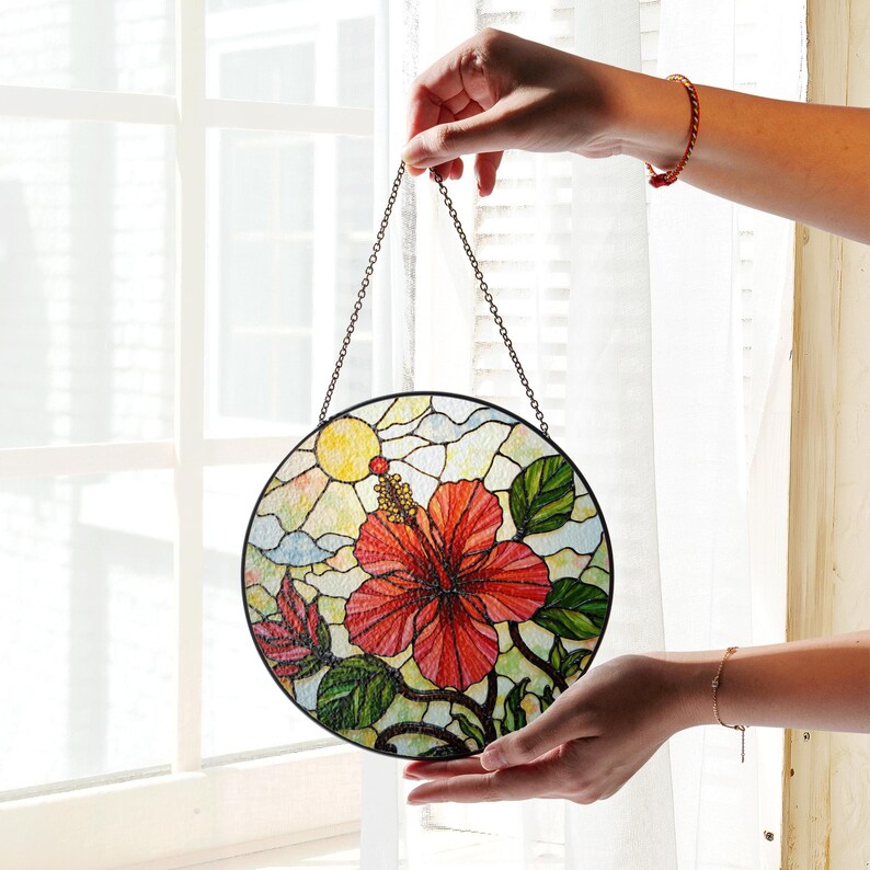 A person's hands holding a round stained glass piece with a vibrant floral design, including a red flower and green leaves, against a window backdrop.
