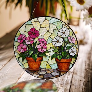 May include: Round stained glass window art featuring two terracotta pots with pink and white flowers. The flowers have green leaves and are set against a colorful, mosaic-style background. A decorative chain is attached for hanging.