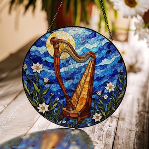 Puede incluir: Una ventana de vitral que representa un arpa con un esquema de color azul, amarillo y marrón. El arpa está rodeada de flores blancas y hojas verdes. La ventana es redonda y tiene una cadena colgando de la parte superior.