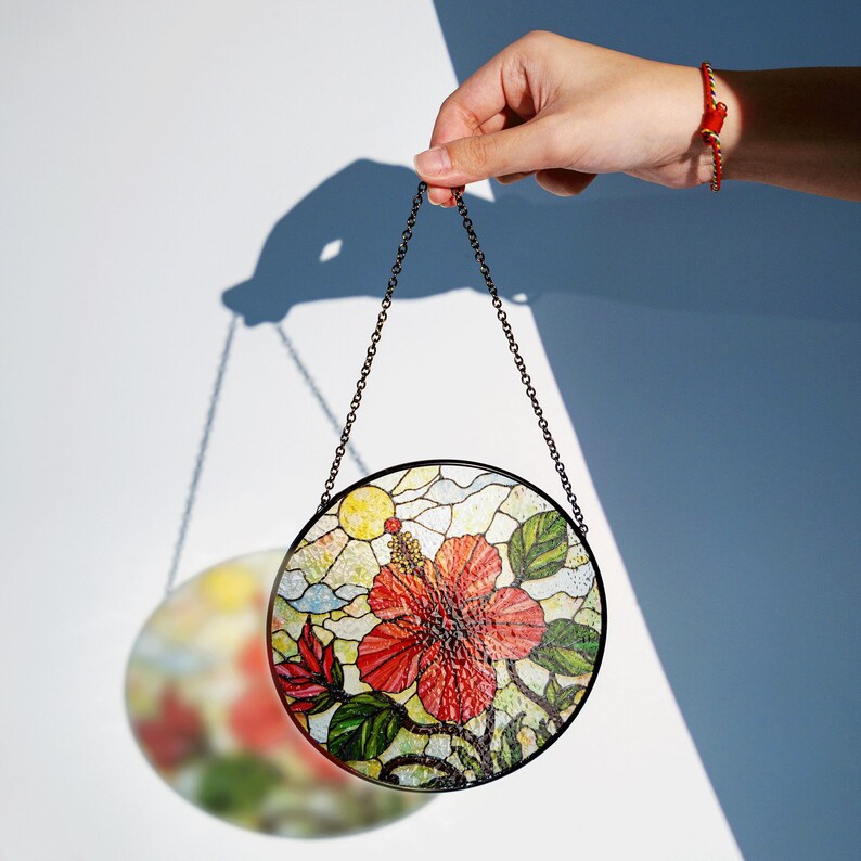 A hand holding a colorful stained glass piece with a floral design, against a white background.