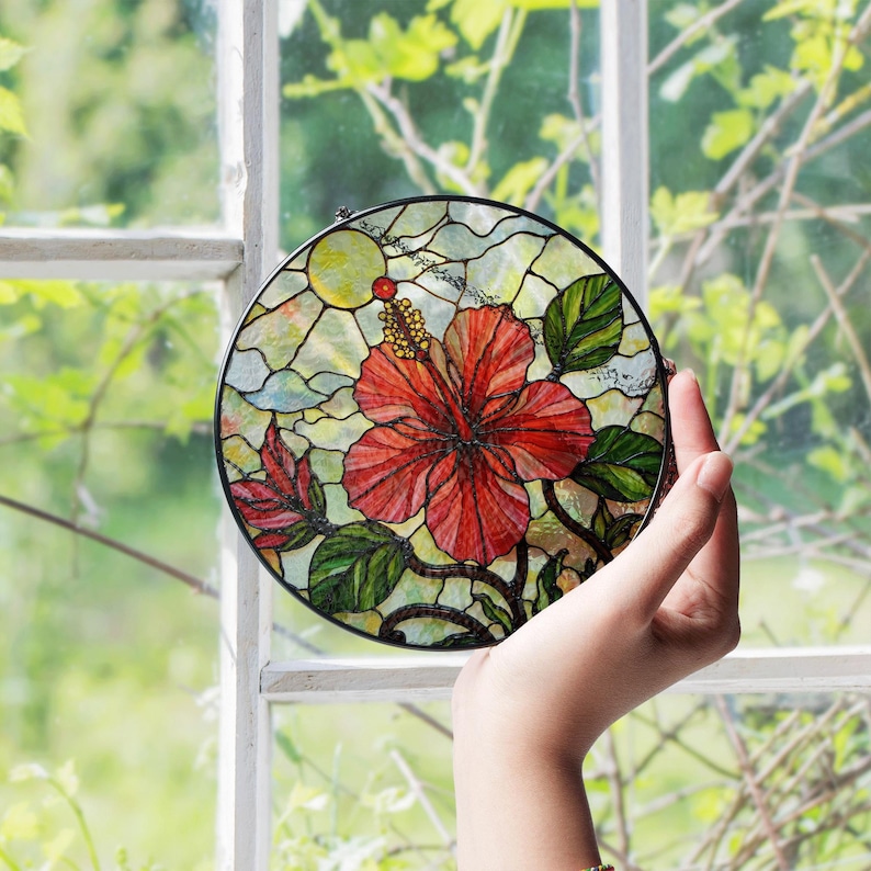 A hand is holding a stained glass piece featuring a vibrant red hibiscus flower against a backdrop of lush greenery.
