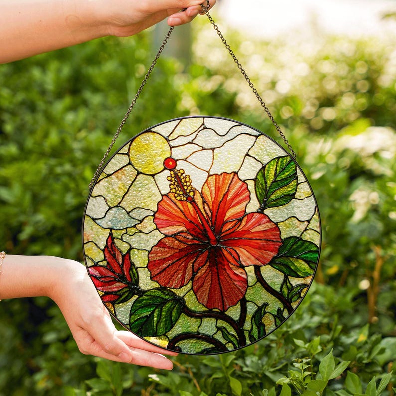 A hand holds a round stained glass piece featuring a vibrant red hibiscus flower surrounded by green leaves against a backdrop of lush greenery.