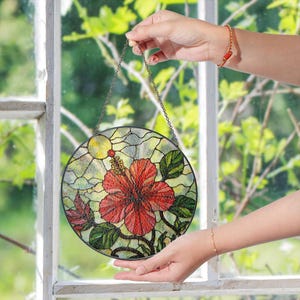 A person's hands holding a colorful stained glass piece with a red flower and green leaves against a blurred green background.
