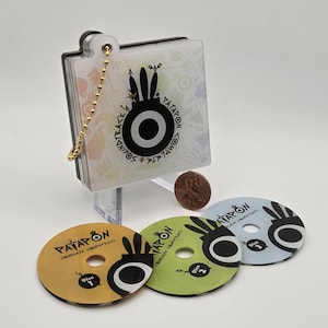 May include: A clear case with a gold chain holds a CD set. Three colorful CDs are displayed below. The CDs are labelled "Disc 1", "Disc 2", and "Disc 3". The case and CDs feature a black and white graphic design with the word "PATAPON".