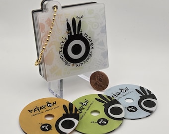 May include: A clear case with a gold chain holds a CD set. Three colorful CDs are displayed below. The CDs are labeled "Disc 1", "Disc 2", and "Disc 3". The case and CDs feature a black and white graphic design with the word "PATAPON".