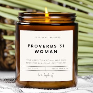 Puede incluir: Un tarro de vela de vidrio marrón con una etiqueta blanca que dice "Let There Be Laughs" y "Proverbs 31 Woman". La etiqueta también incluye el texto "Some light for a woman who rises before the sun, or at least tries to." y "9 oz / 2550 Vegan, Made in USA live. laugh. lit"