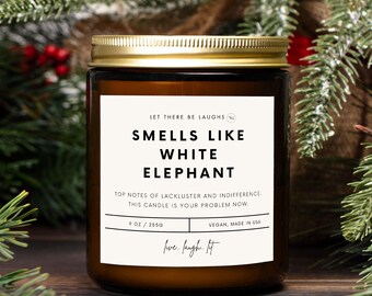 Könnte beinhalten: Braune Glas-Kerze mit goldenem Deckel und weißem Etikett. Auf dem Etikett steht "Smells Like White Elephant" mit dem Text "Let There Be Laughs" darüber. Die Kerze hat 255 g.