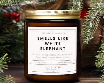 Könnte beinhalten: Eine braune Glas-Kerze mit goldenem Deckel. Das Etikett lautet "SMELLS LIKE WHITE ELEPHANT" mit dem Text "TOP NOTES OF LACKLUSTER AND INDIFFERENCE. THIS CANDLE IS YOUR PROBLEM NOW." Die Kerze ist 255g und vegan.